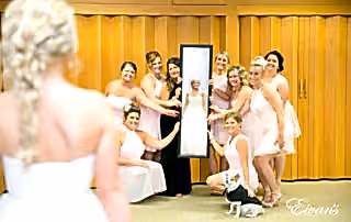 The photographer captures a reflective photo of a bride smiling and looking into a rectangular mirror held by her excited bridal party. Her small, tuxedo-clad dog sits sweetly alongside the bridesmaids.