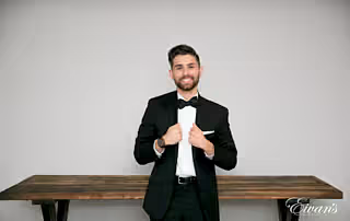 The groom grips the lapels of his tuxedo jacket and smiles broadly in front of a vintage wood table.