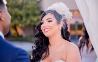 New Bride smiles adoringly at her new husband during the ceremony. The Bride wears an ivory, feathered birdcage visor with netting.