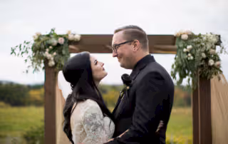 Rustic wedding photography. Bride and Groom embrace each other beneath a florally decorated atrium in an open field of wildflowers on an overcast day.