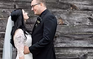 Bride and Groom look loving at one another posed in front of a rustic wooden cabin. The Groom is wearing a black rose boutonniere.
