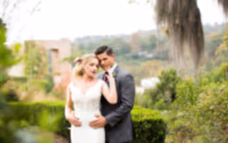 Bride and Groom give the photographer a sultry glance while in the garden. Greenery and old Willow trees rest in the background complimenting the Bride's floral hairpiece with deep red accents.